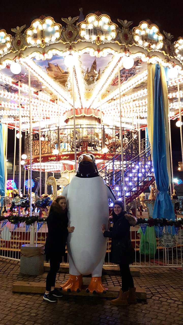 Foto: Zwei Studierende lehnen an einer übergroßen Pinguinfigur. Dahinter ein großes Kinderkarussell.