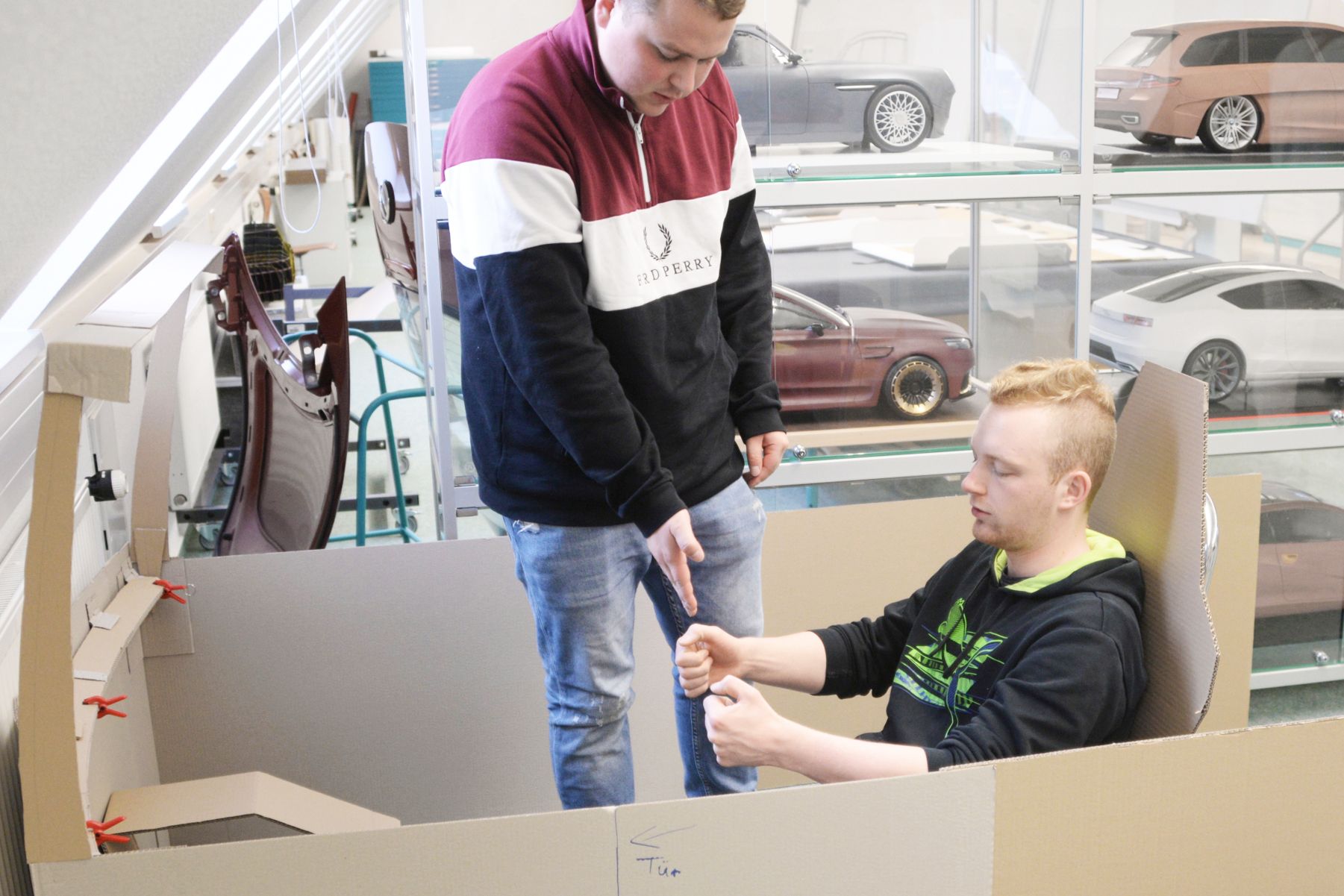 Foto: Zwei Studierende (einer sitzt, der andere steht) in einem KFZ-Modell aus Pappe.