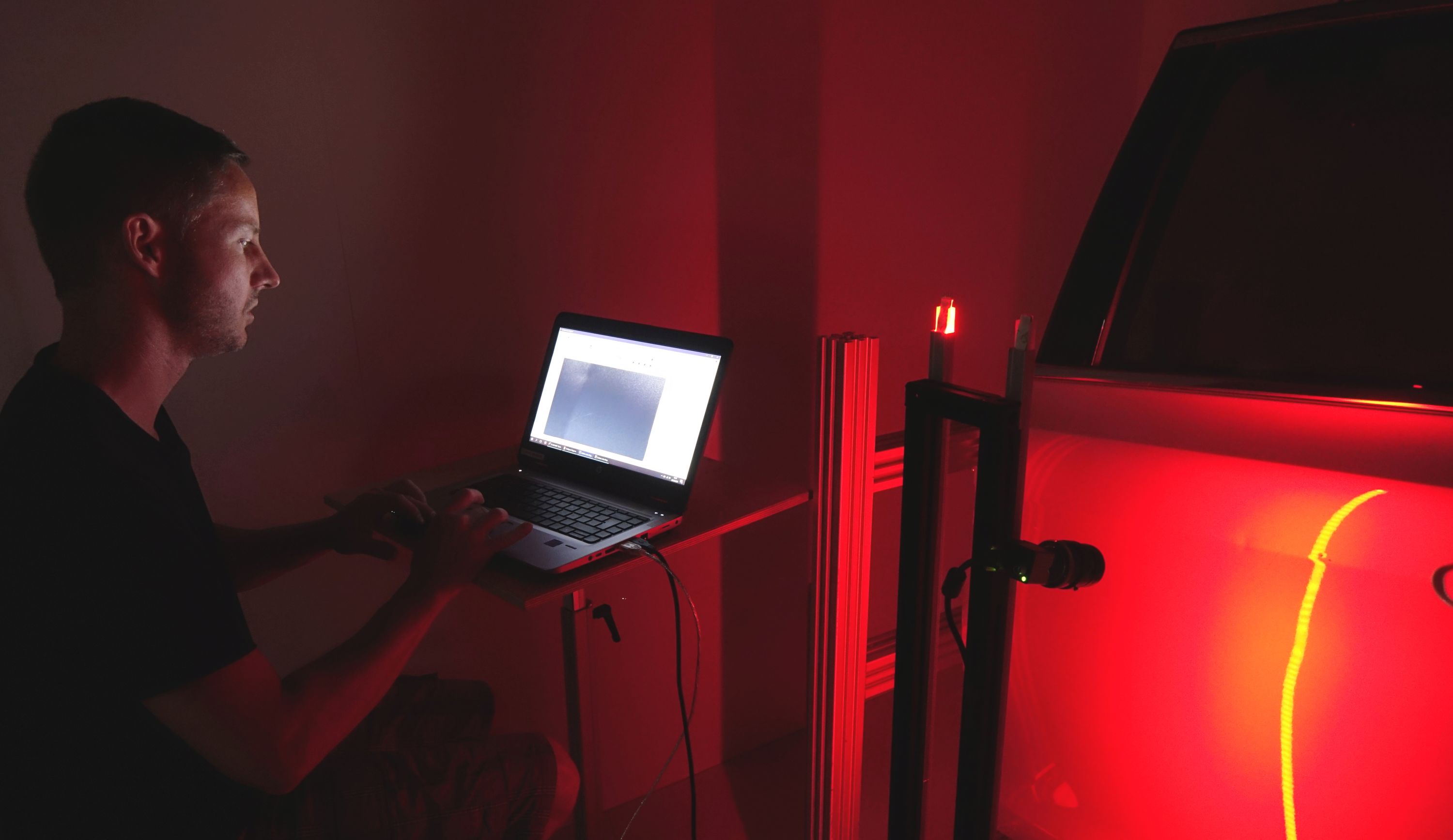 Foto: Ein Mann sitzt im Dunkeln vor einem Laptop. Daneben ist ein in rot getauchtes Fahrzeug zu sehen. 