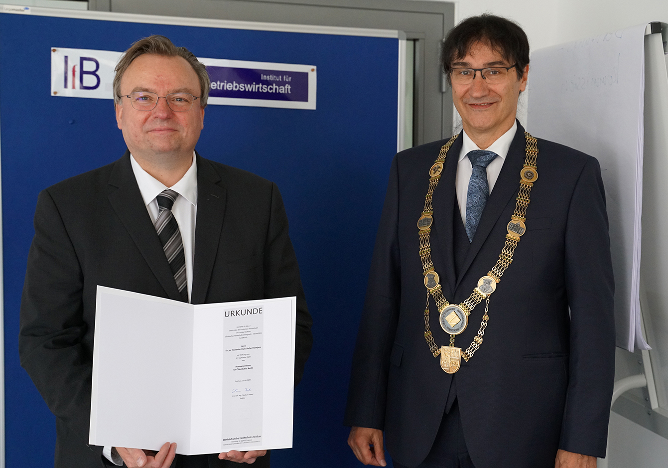 Foto: Prof. Haentjens links mit der Urkunde zur Bestellung als Honorarprofessor. Rechts neben ihm Rektor Prof. Stephan Kassel mit Amtskette. 