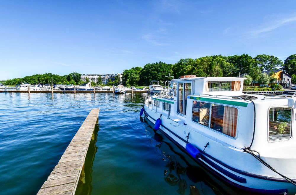 Foto: Im Hintergrund ist ein kleiner, naturnaher Hafen. Rechts vorn liegt ein Motorboot, in der Mitte des Bildes ist ein Holzsteg, der ins Wasser reicht. 
