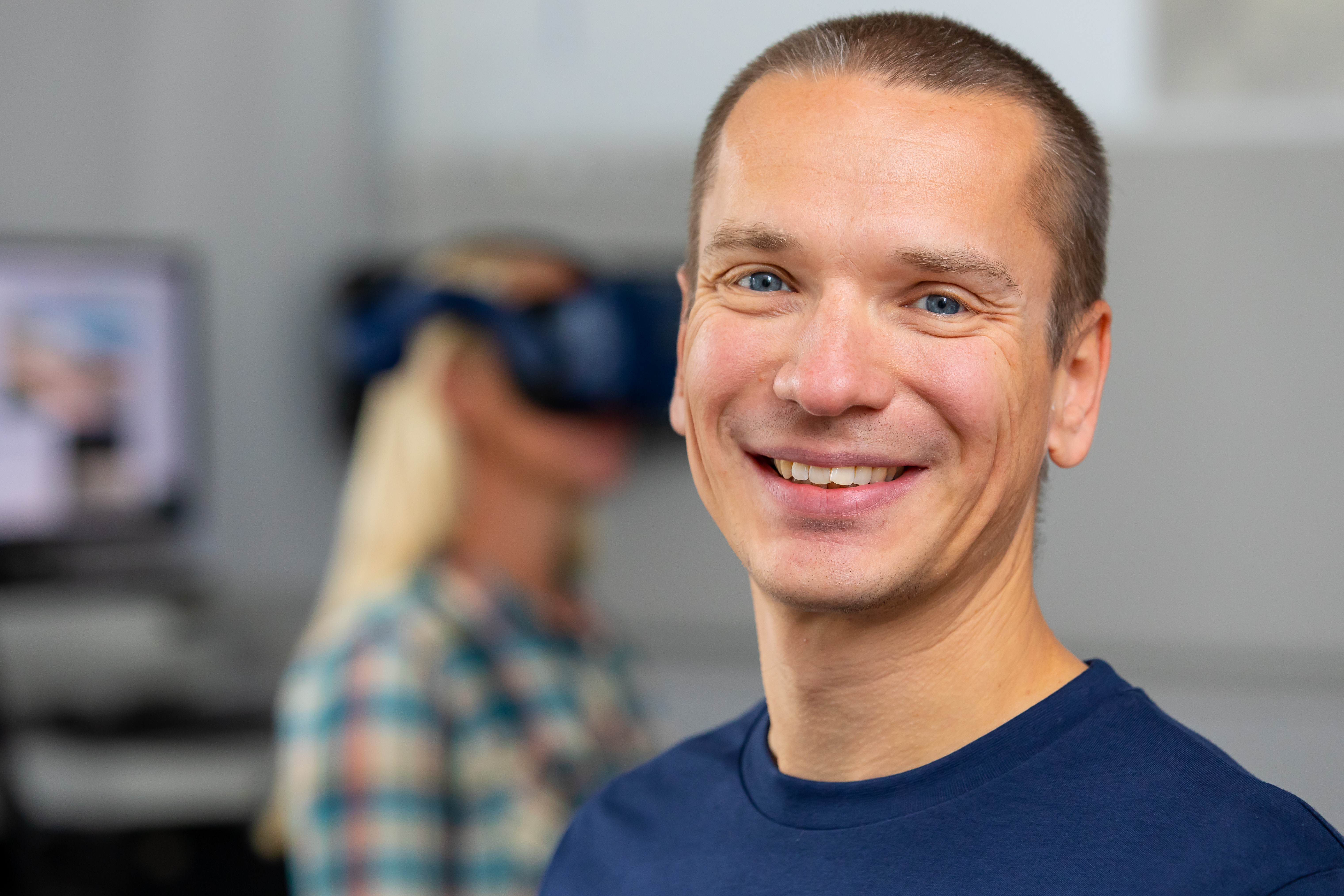 Foto: Prof. Sven Hellbach blickt lächelnd in die Kamera. Er trägt ein blaues T-Shirt. Im Hintergrund ist verschwommen eine Studierende mit einer VR-Brille zu sehen. 
