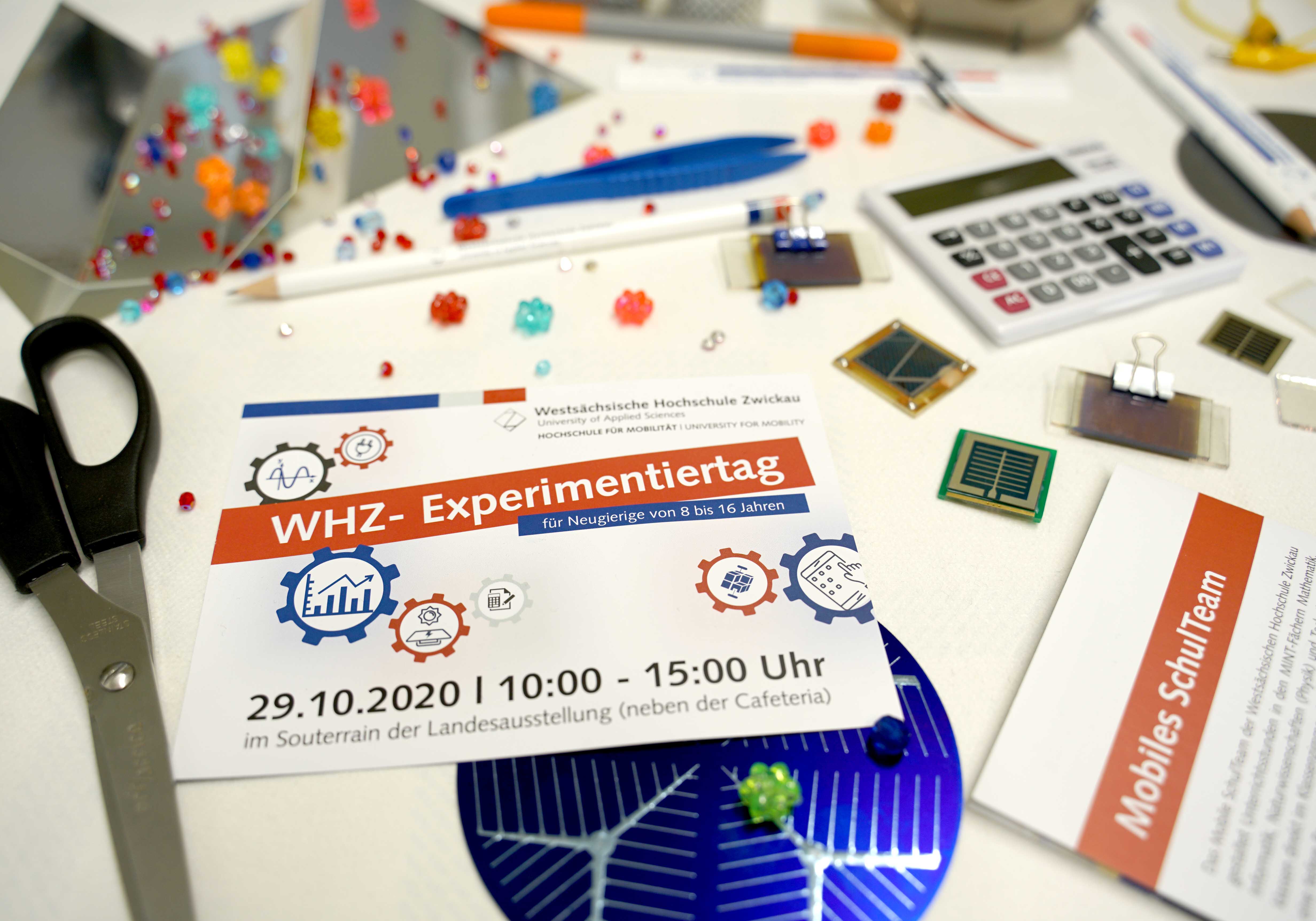 Foto: Diverse Gegenstände (Schere, Taschenrechner, Mikrochips, Stifte) liegen auf einem Schreibtisch. In der Mitte ein Flyer mit er Aufschrift "WHZ-Experimentiertag"
