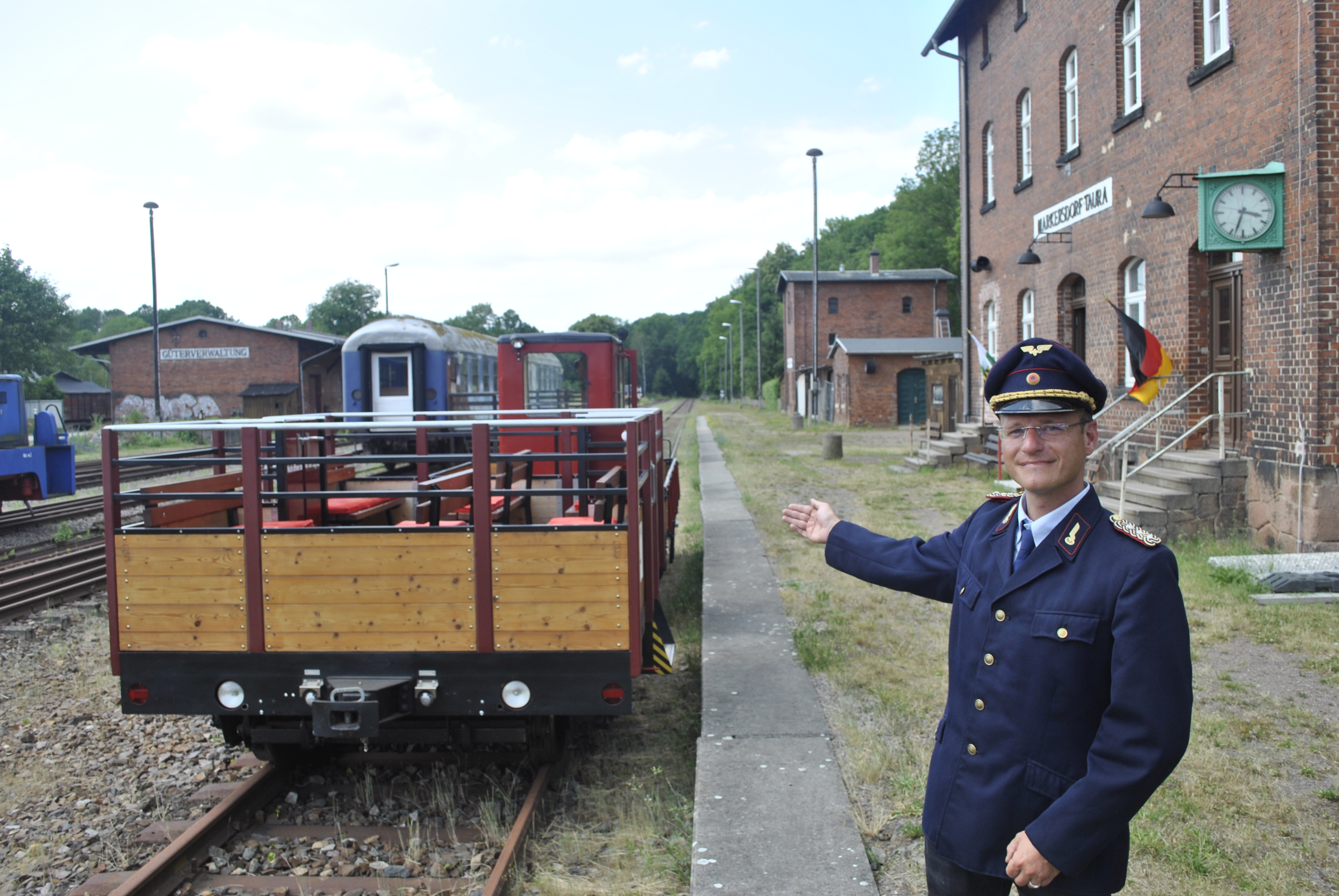 Foto: Hr. Martin Bauch, in Bahnkleidung, steht vor einem historischen Bahnhofsgebäude und zeigt in Richtung eines historischen Personenzuges. 