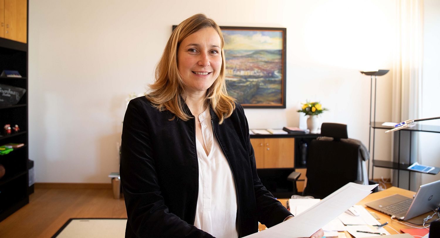 Foto: Eine Frau steht lächelnd in einem Büro und hält Unterlagen in der Hand. 