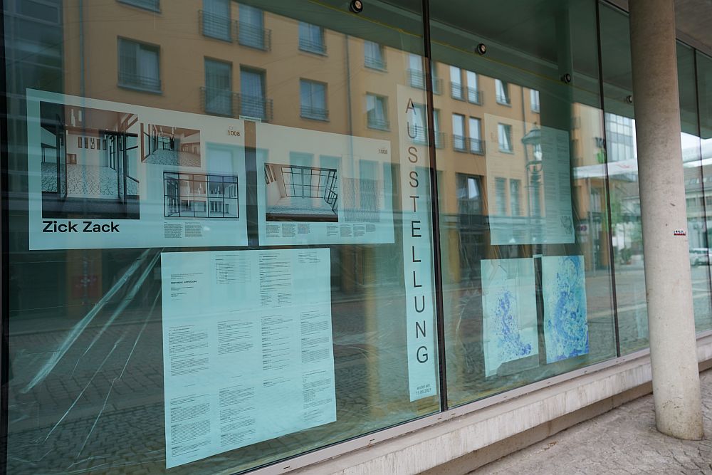 Foto: An den großen Fenstern der Hochschulbibliothek sind eingereichte Entwürfe für den Wettbewerb Kunst am Bau zu sehen. In der Mitte des Bildes ist das Wort Ausstellung zu lesen.