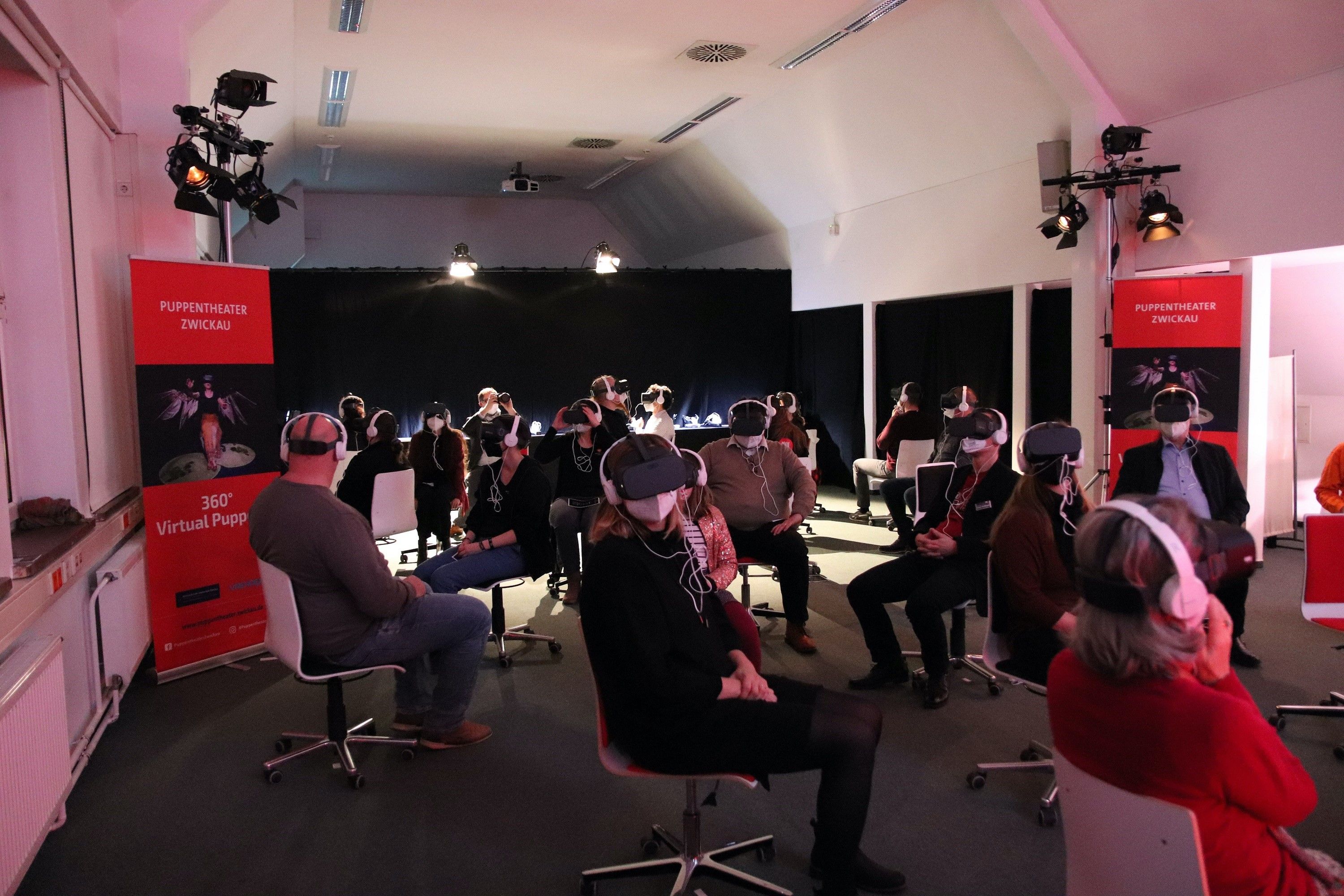 Foto: In einem schwach beleuchteten Raum sitzen viele Menschen mit Abstand auf Drehstühlen und haben alle VR-Brillen und Kopfhörer auf.