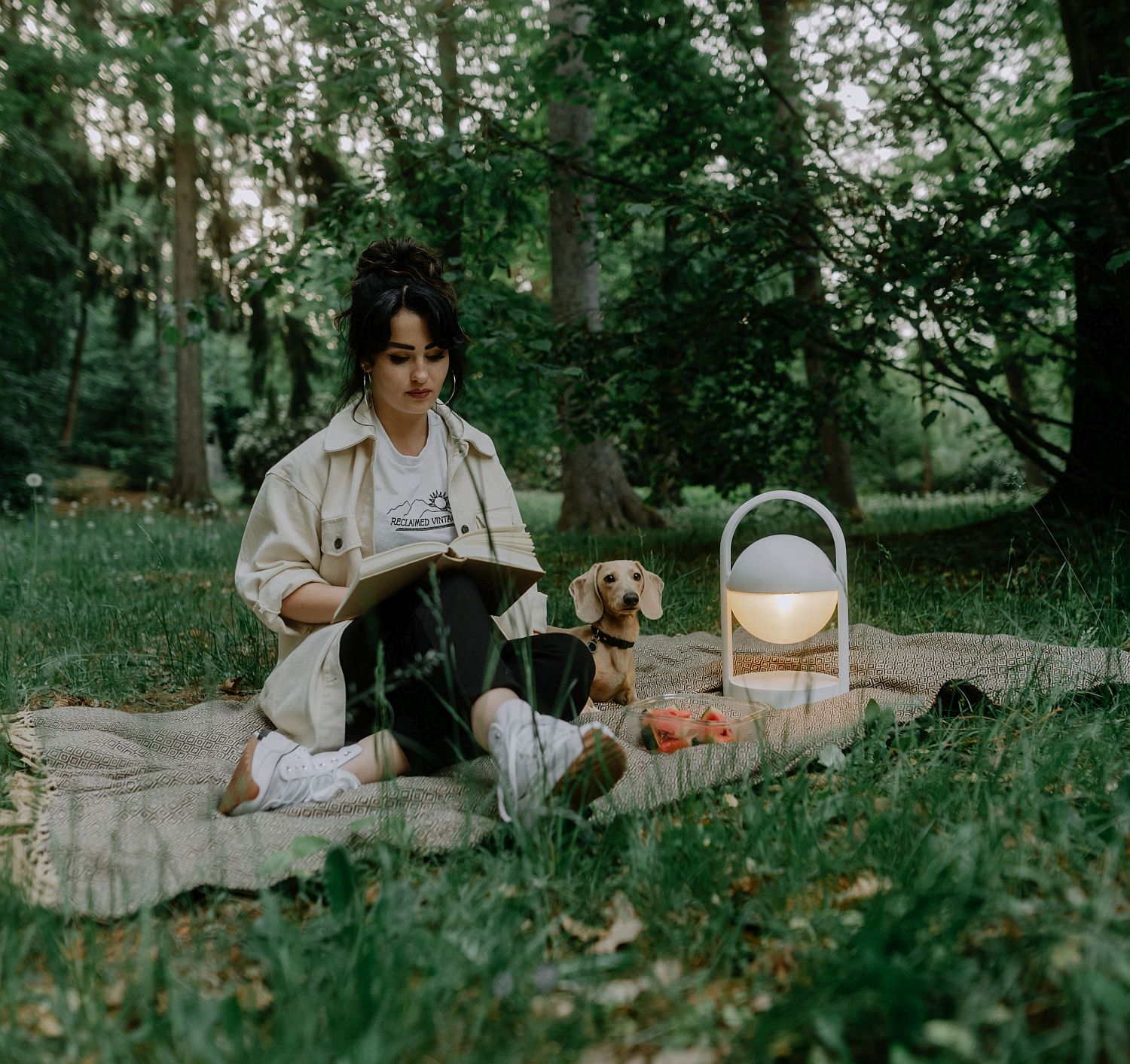 Eine Frau sitzt auf einer Decke in einem Wald und liest. Neben ihr liegt ein Dackel und daneben steht eine modern gestaltete Lampe.