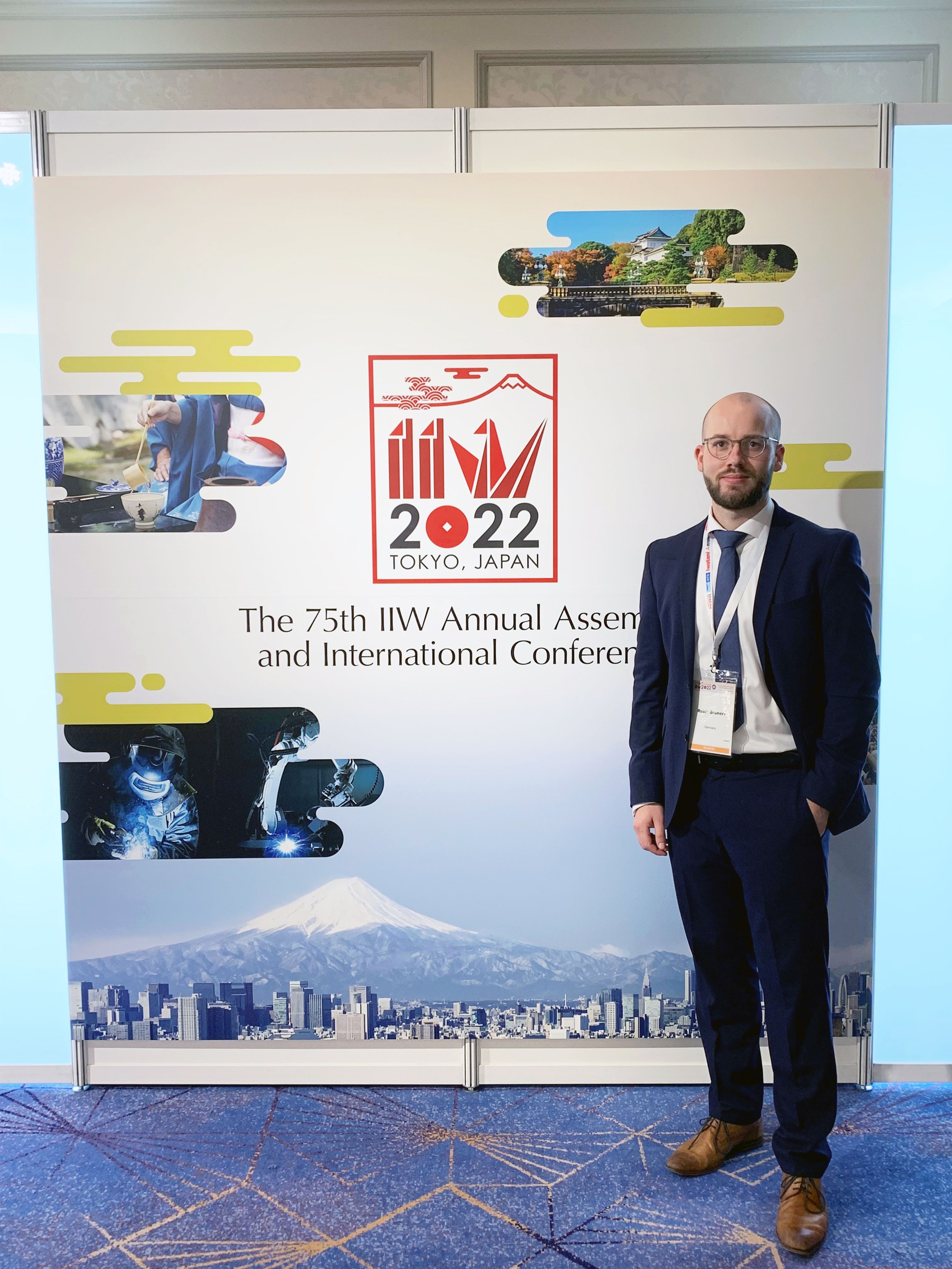 Ein Mann im Anzug steht vür einem Logo der "IIW Annual Assembly and International Conference on Welding and Joining", Tokyo 2022
