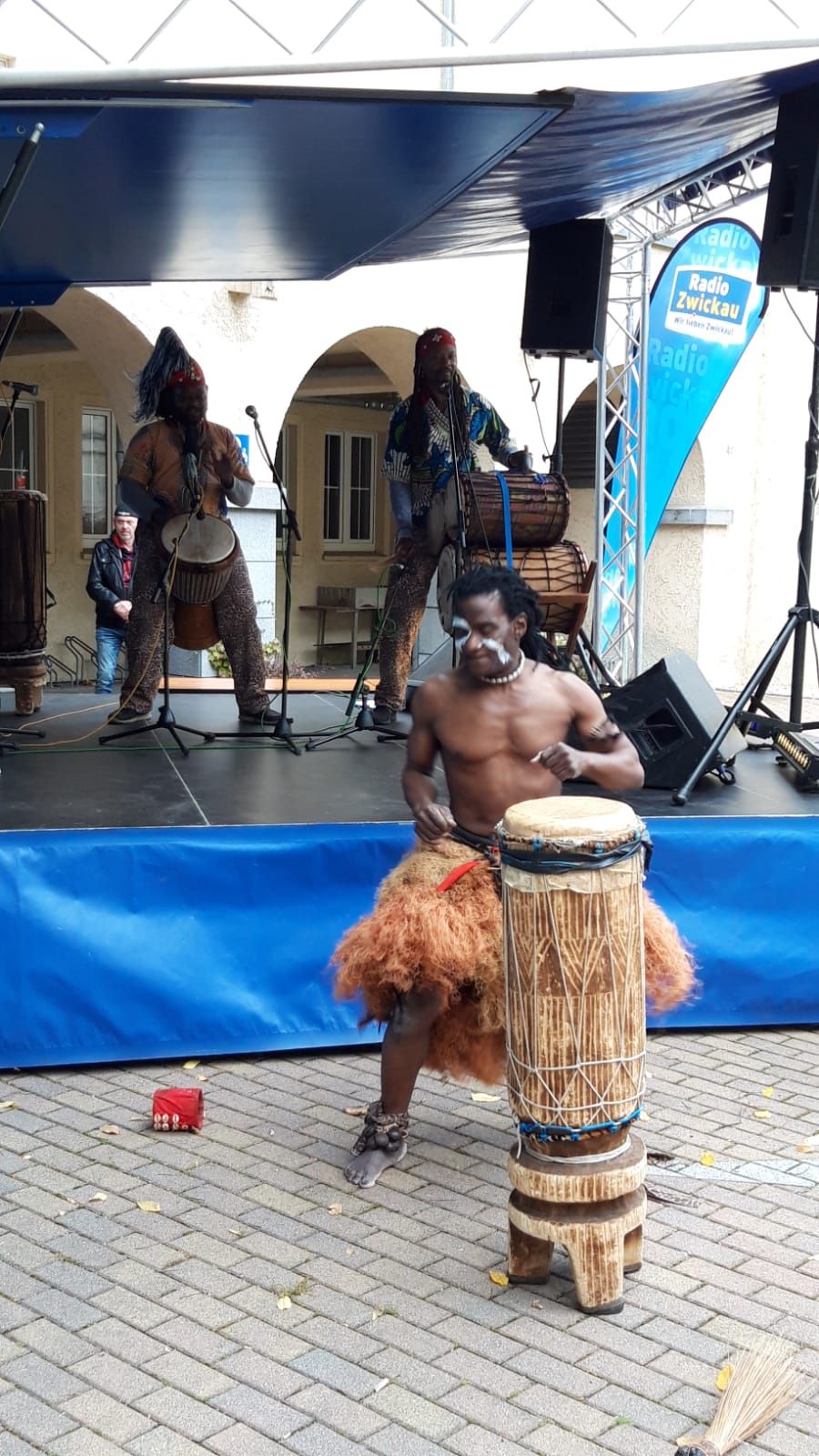 Ein afrikanischer Musiker steht auf einem gepflasterten Platz hinter einer traditionellen afrikanischen Trommel. Dahinter stehen auf einer Bühne zwei weitere Männer mit verschiedenen Trommelinstrumenten. Alle tragen traditionelle afrikanische Kleidung. 