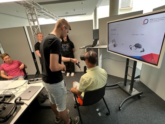 Personen sitzen in einem Labor und arbeiten mit einer VR-Brille. Im Hintergrund ist eine digitale Leinwand zu sehen. 