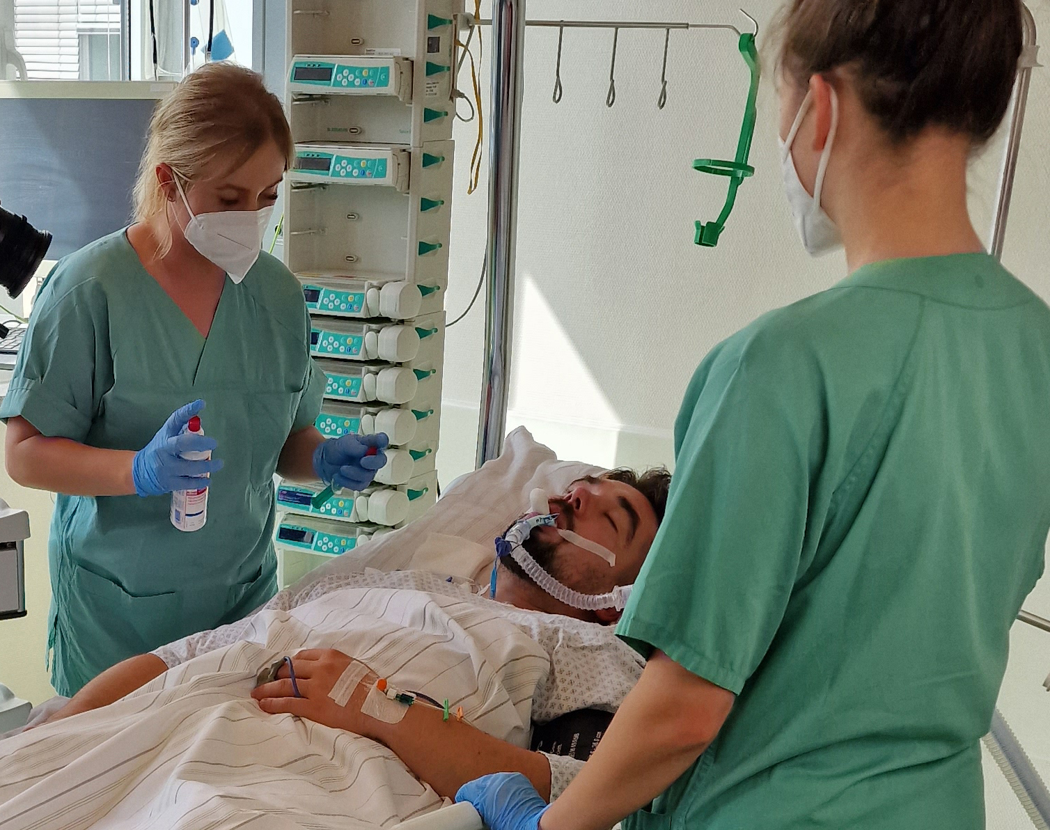 Ein Patient liegt im Bett und wird beatmet. Zwei Schwestern mit Mund-Nasenmasken betreuen ihn. 