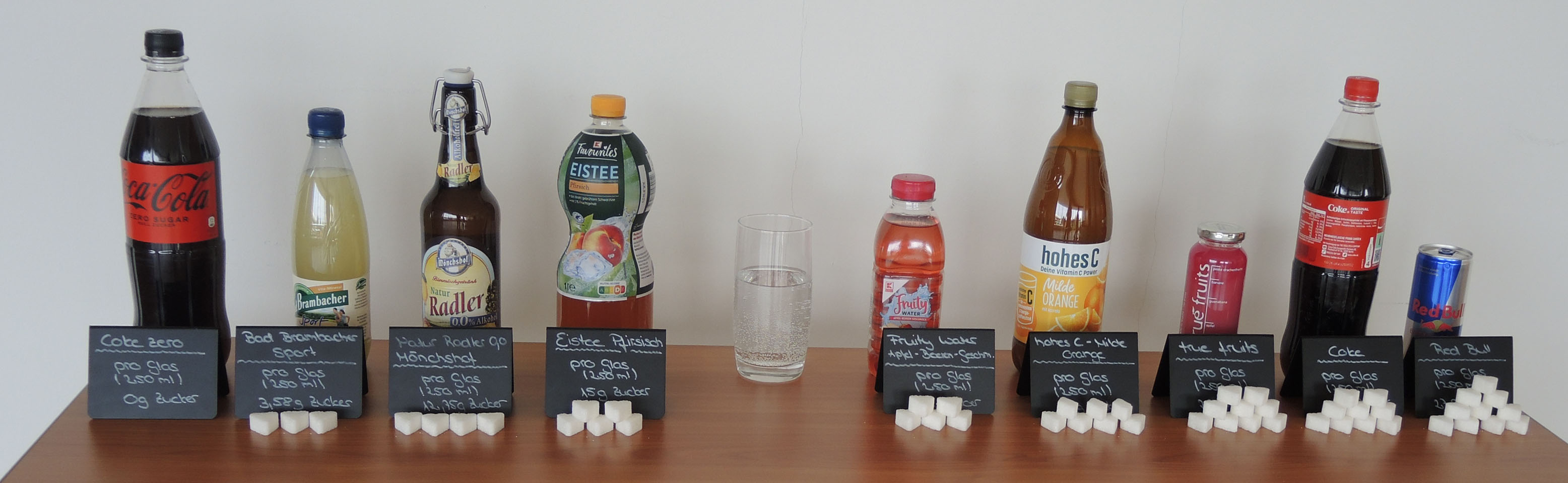 Flaschen mit verschiedenen Getränken stehen auf einem Tisch, Zuckerwürfel vor den Getränken zeigen den Zuckergehalt an. 