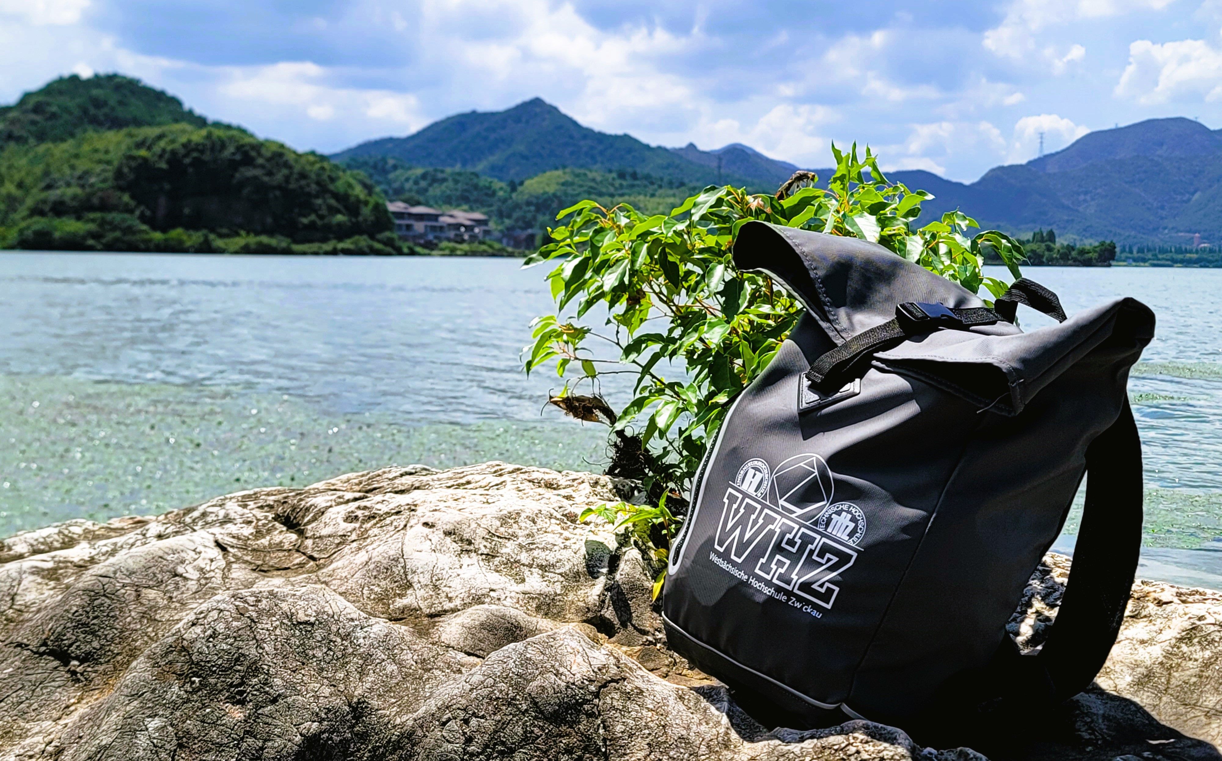 Ein Rucksack mit WHZ-Logo steht an einem Strand, im Hintergrund ein klarer See und Berge