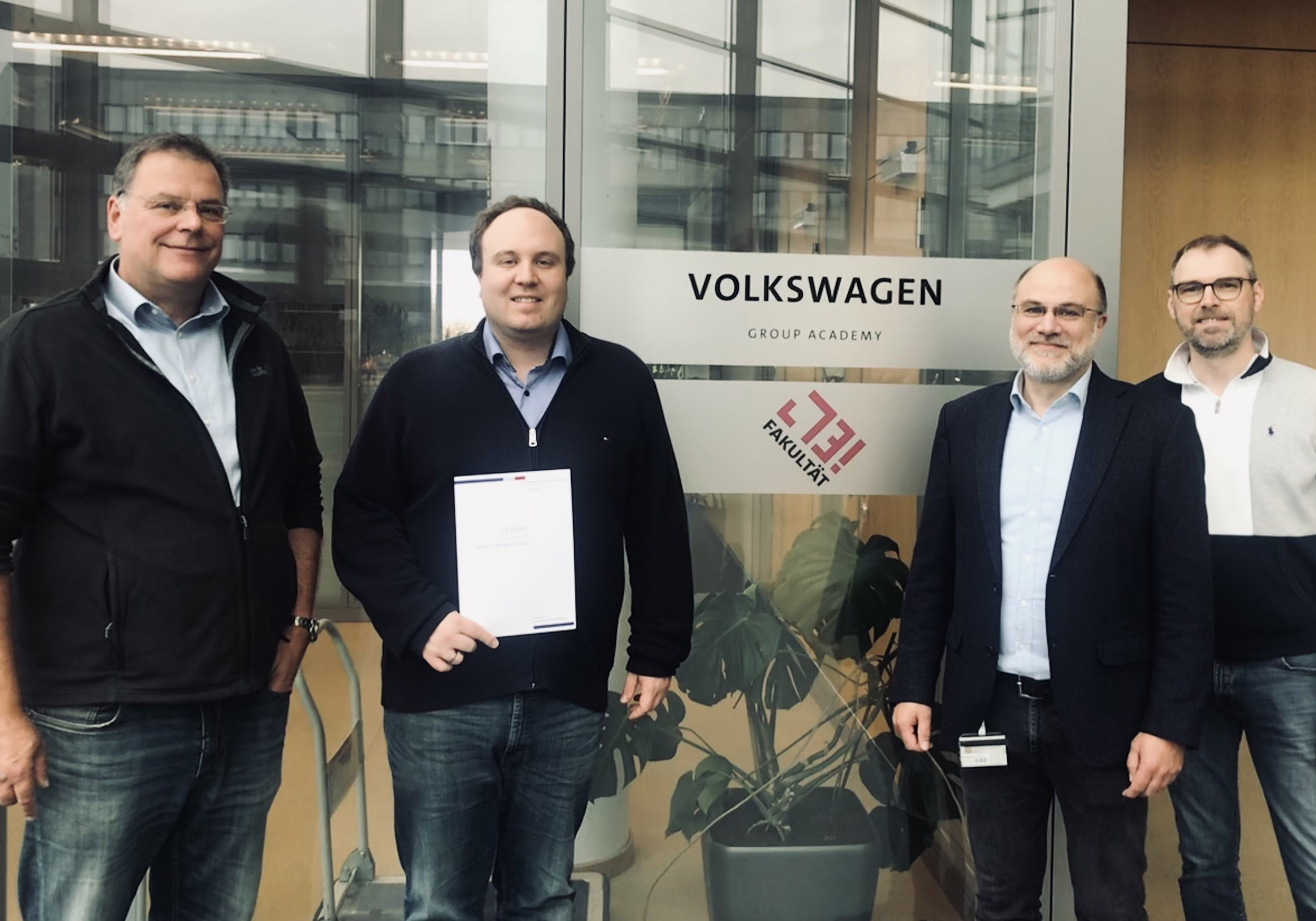 Gruppenbild von vier Personen, eine hält eine Urkunde in der Hand, im Hintergrund der Schriftzug Volkswagen Group Academy. 