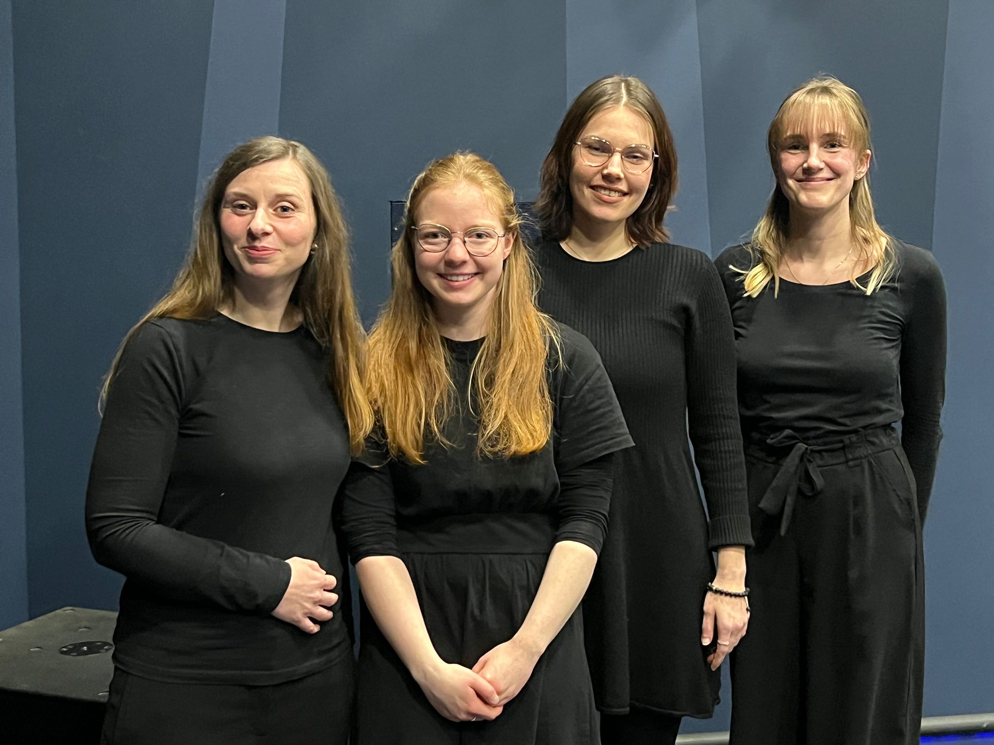 Vier Frauen in schwarzer Kleidung, die nebeneinander stehen und freundlich nach vorn schauen