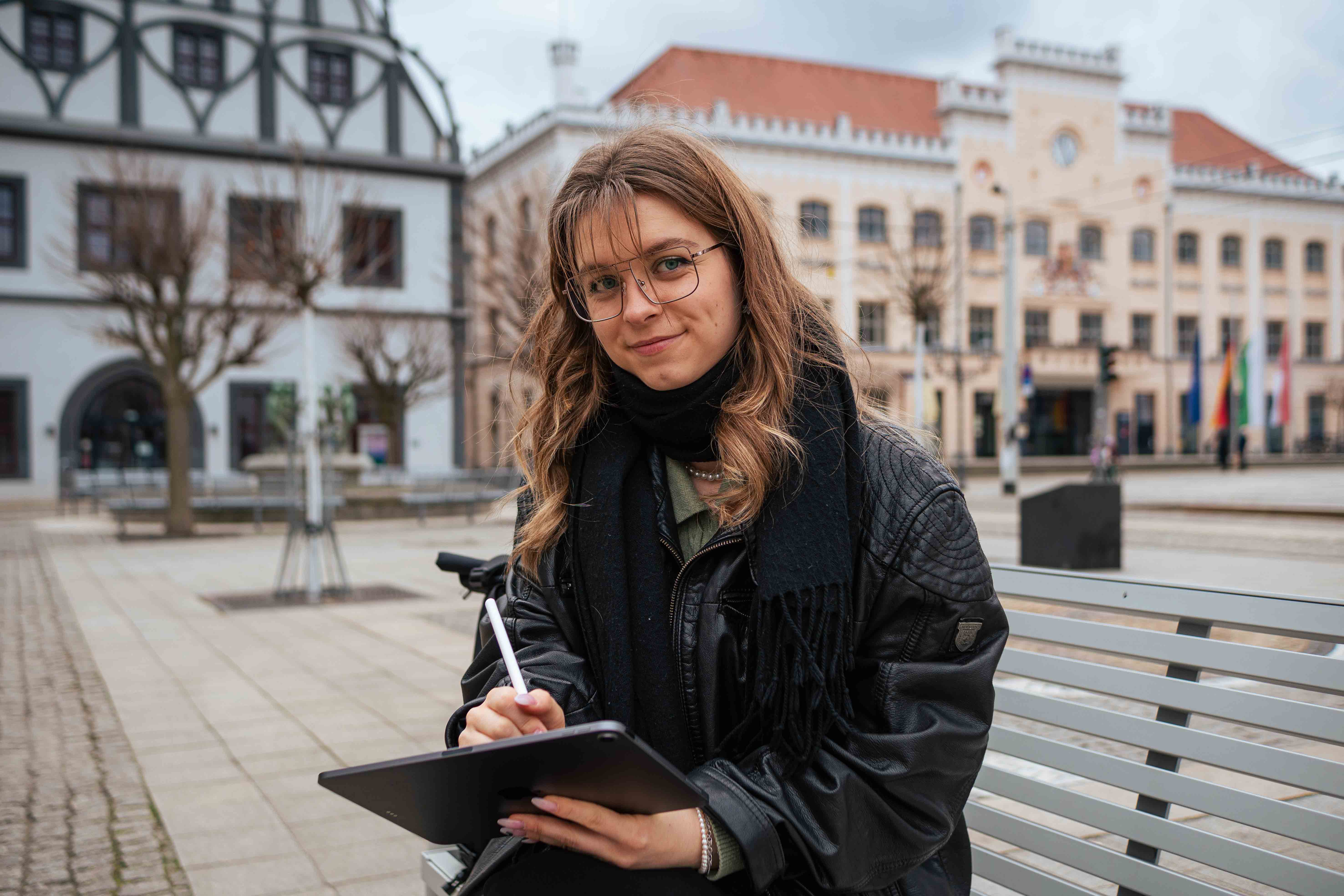 Eine Studentin sitzt mit Tablet auf dem Zwickauer Hauptmarkt und blickt in die Kamera