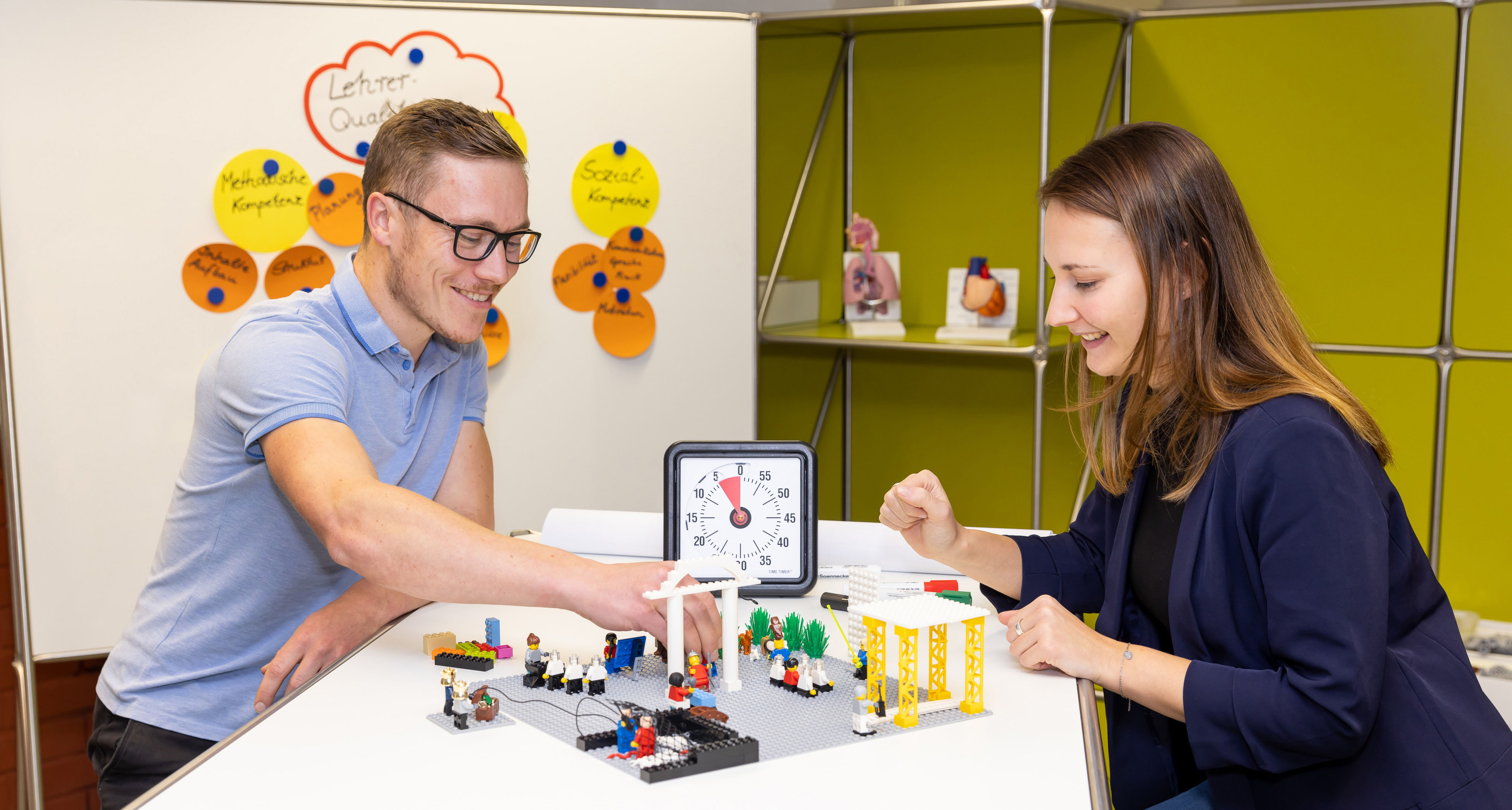 Zwei Studierende sitzen an einem Tisch und stellen Abläufe in einem Krankenhaus mit Lego-Bausteinen dar.
