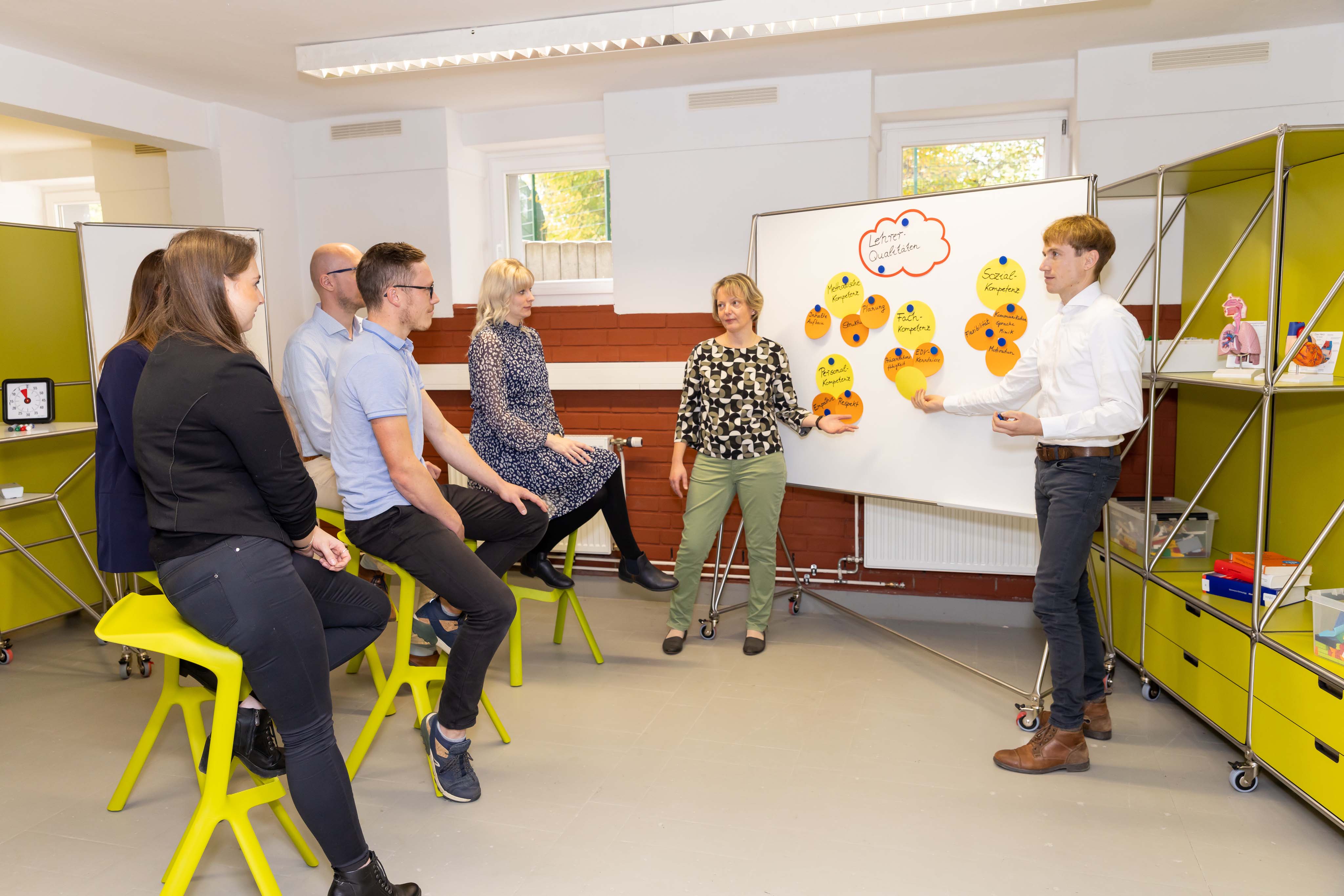 Zwei Personen erklären an einer Tafel, anderer Personen sitzen auf Sitzhockern davor und hören zu.